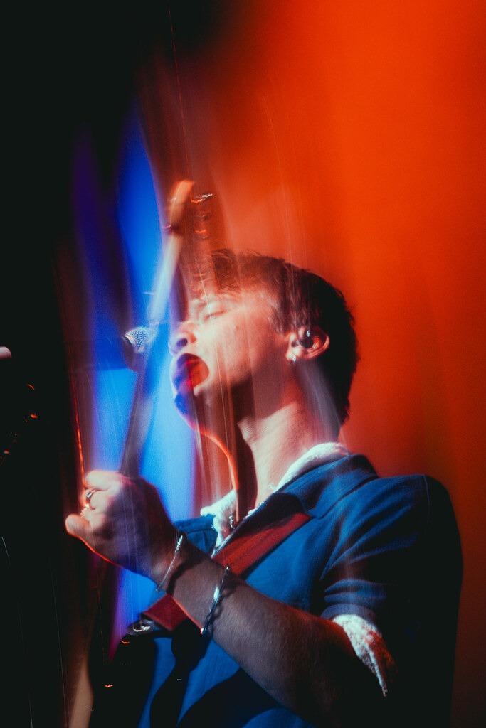 A Balu Brigada musician sings into a microphone in red and blue stage lighting.