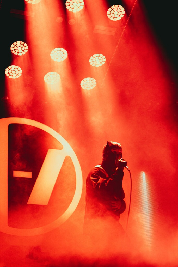 A masked singer stands on stage beneath red spotlights and haze.
