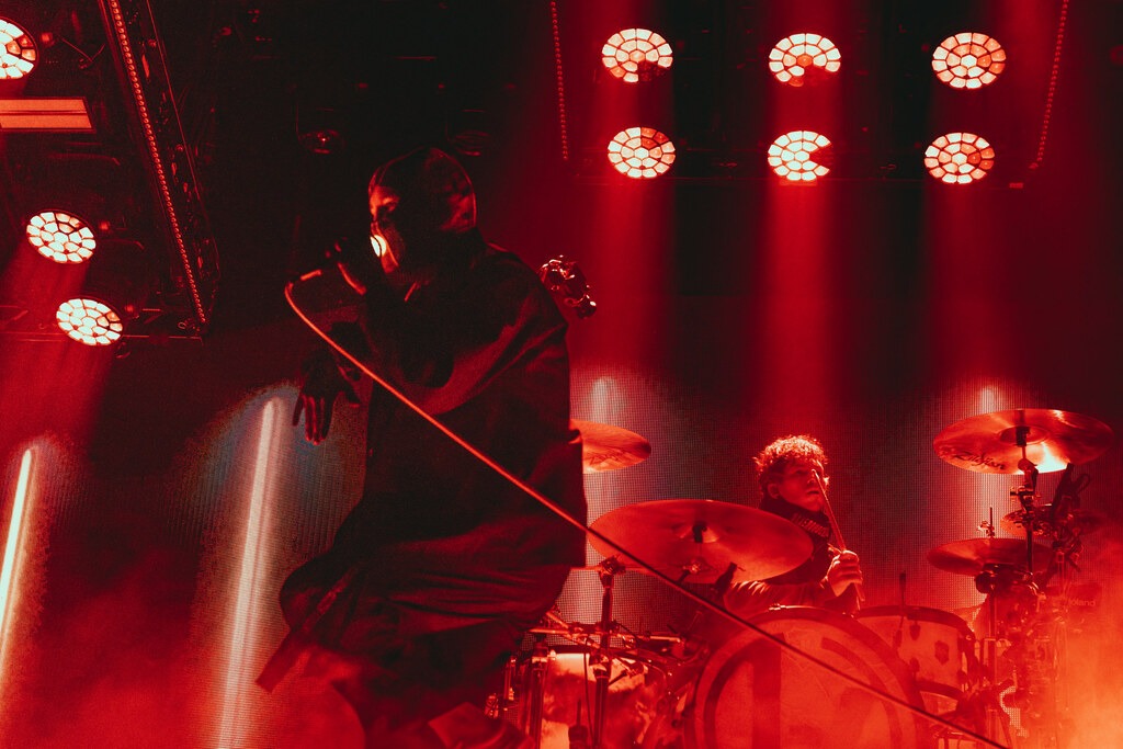 Twenty One Pilots perform under red stage lights, with a singer in the foreground and the drummer behind.