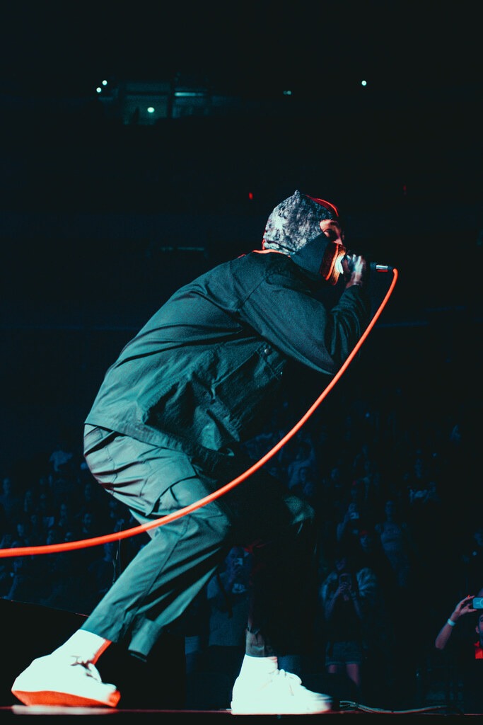 A Twenty One Pilots performer crouches at the edge of the stage with a microphone.