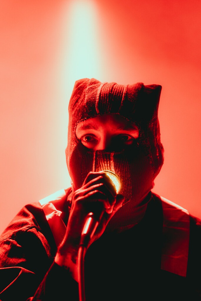 Close-up of a masked Twenty One Pilots singer holding a microphone beneath a red spotlight.